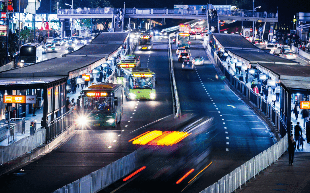 Nighttime urban bus terminal with multiple buses at platforms and a pedestrian bridge overhead, motion blur on moving vehicles, crowd on sidewalks.